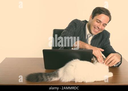 Un homme d'affaires persan souriant et touchant un chat mignon tout en étant assis avec un ordinateur portable sur une table en bois Banque D'Images