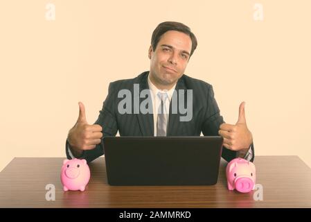Homme d'affaires persan donnant des pouces avec ordinateur portable et deux banques de piggie sur table en bois Banque D'Images