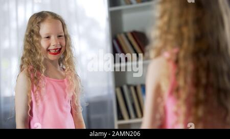 Petite fille tachée de rouge à lèvres riant dans le miroir, appliquant le premier maquillage Banque D'Images