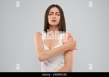 Young caucasian woman in white shirt posant sur fond gris de toucher l'épaule avec isolés expression douloureuse à cause de blessures. Vue avant de brune aux yeux clos et bouche ouverte. Banque D'Images