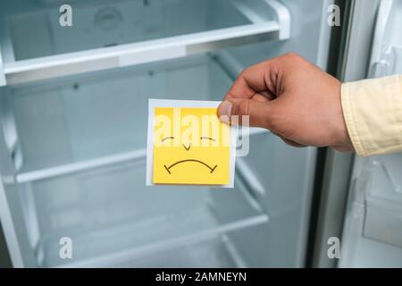 vue réduite de l'homme tenant un smiley triste près d'un réfrigérateur vide ouvert Banque D'Images