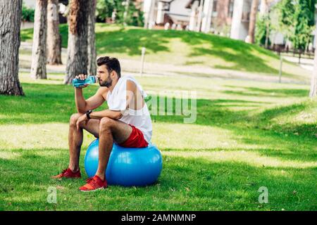 un sportif barbu buvant de l'eau de la bouteille de sport tout en étant assis sur le ballon de fitness Banque D'Images