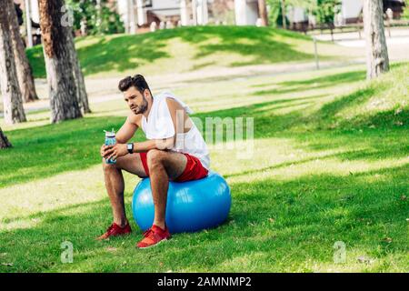 sportif barbu tenant une bouteille de sport tout en étant assis sur le ballon de fitness Banque D'Images