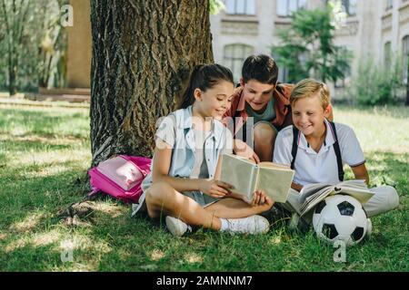 trois camarades de classe mignons lisent le livre et sourient tout en s'asseyant sur la pelouse sous l'arbre Banque D'Images