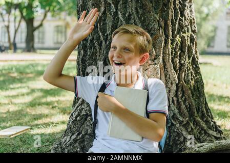 un écolier gai avec un livre qui agait la main tout en étant assis sous l'arbre et en regardant loin Banque D'Images