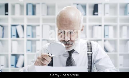 Homme d'affaires senior lisant la paperasserie à l'aide d'une loupe, il vérifie soigneusement un accord Banque D'Images