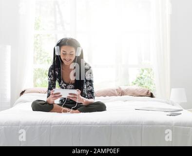 Fille se reposant dans sa chambre, elle est assise sur le lit, regarder des vidéos et écouter de la musique en ligne avec sa tablette Banque D'Images