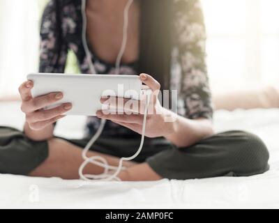 Fille se reposant dans sa chambre, elle est assise sur le lit, regarder des vidéos et écouter de la musique en ligne avec sa tablette Banque D'Images