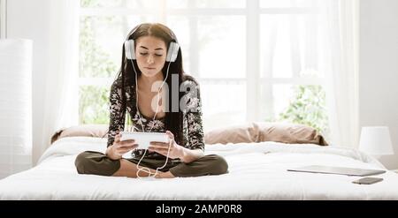 Fille se reposant dans sa chambre, elle est assise sur le lit, regarder des vidéos et écouter de la musique en ligne avec sa tablette Banque D'Images