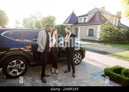 Multiethnical avec succès trois affaires confiant dans smart casual wear à l'aide de tablet près de voiture, debout à l'extérieur dans la cour de l'entreprise moderne Banque D'Images