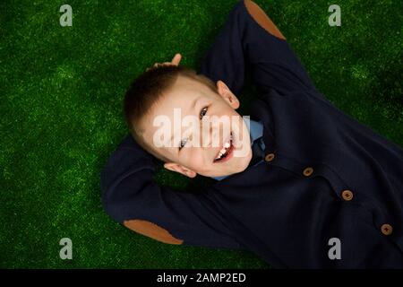 Un petit garçon s'allonge sur le dos, regardant la caméra et souriant.. Vue de dessus d'un mignon petit garçon heureux looking at camera et souriant alors que se trouve sur une pelouse Banque D'Images