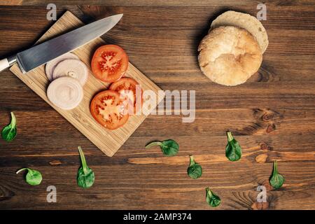 couteau avec des tranches de tomate et d'oignon sur une planche à découper à côté d'un pain et quelques feuilles vertes sur une table en bois, vue sur le dessus, la nourriture végétalienne et la hea Banque D'Images