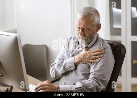 Inquiet homme senior ayant une crise cardiaque, il est assis au bureau et touche sa poitrine Banque D'Images