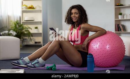 Femme biraciale dans une salle de gym assis au sol, à l'aide d'une application de téléphone mobile Banque D'Images