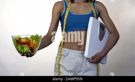 Femme en pantalon ample debout avec pèse-personne, tenant hors bol de salade, régime Banque D'Images