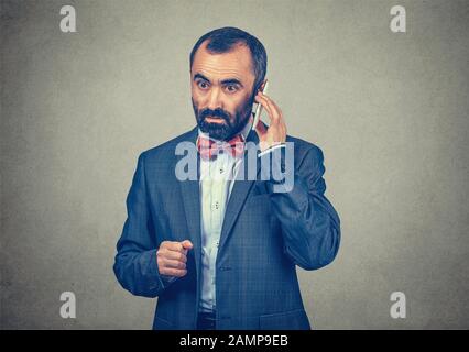 Homme d'affaires parlant sur un téléphone mobile écoutant ses messages vocaux avec une expression sérieuse sur le visage. Modèle mixte barbu isolé sur gre gris Banque D'Images