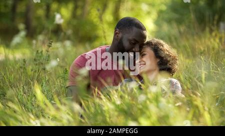 Couple multiracial riant et chat tout en couché dans l'herbe, date extérieure Banque D'Images
