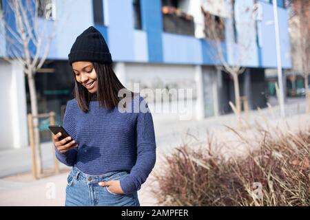 Portrait de la jeune femme africaine américaine portant une casquette en laine debout dans la rue tout en utilisant un téléphone portable Banque D'Images