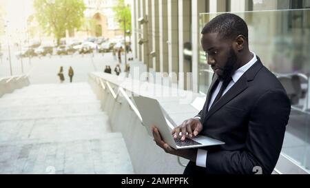 Homme d'affaires noir qui vérifie les fichiers sur l'ordinateur portable, en surtravaillant pendant la pause déjeuner Banque D'Images