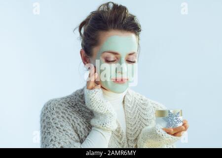 cette élégante femme présente un sweat-shirt à col roulé et un gilet avec masque facial vert qui touche le visage et tient un bol cosmétique et un flocon de neige sur le dos bleu clair d'hiver Banque D'Images