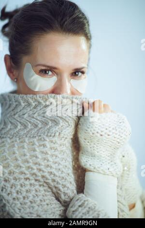 Portrait d'une femme élégante dans un sweat-shirt à col roulé et un gilet avec des taches oculaires cosmétiques dans le cadre des soins de la peau d'hiver se cachant derrière des vêtements contre la neige Banque D'Images