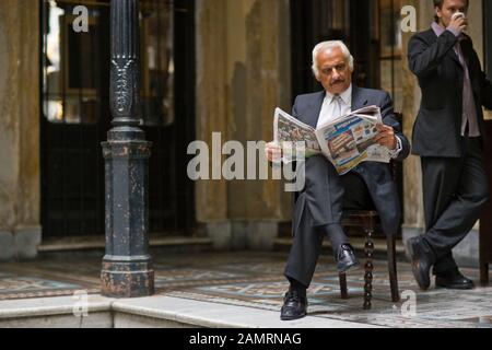 Homme adulte mûr assis sur une chaise lisant un journal alors qu'un jeune homme boit à côté de lui. Banque D'Images