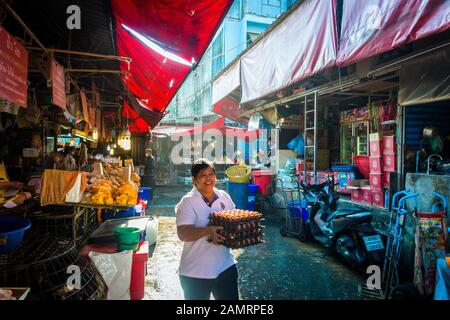 Marché de Khlong Toei à Bangkok, Thaïlande avec des étals de nourriture sous un toit rouge avec une femme tenant des caisses d'œufs passant par la rue étroite Banque D'Images