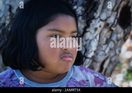 Une petite fille avec une expression faciale bouleversée se tenant devant un arbre. Banque D'Images