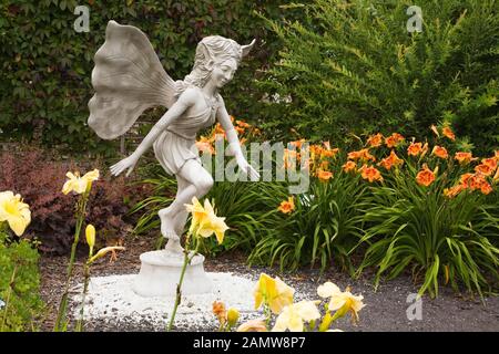 Statue d'une fée mythique à ailes bordées de jaune 'Vanila Fluff' et orange 'Tiger' Hemerocallis - fleurs de jour dans le jardin de fées en été. Banque D'Images