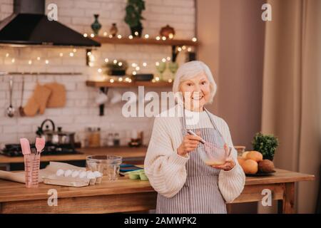 Une dame souriante aux cheveux gris se tenant près de la table dans la cuisine Banque D'Images