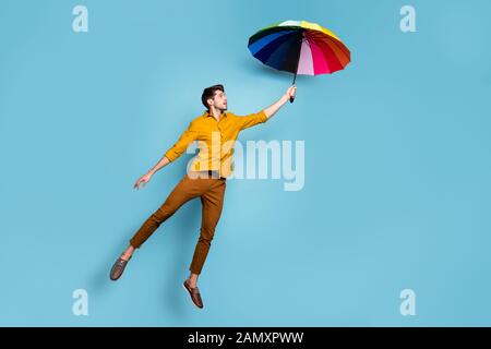 Photo pleine longueur de crazy guy jumping high holding parapluie lumineux colorés battant de plus porter un pantalon chemise jaune fond couleur bleu isolé Banque D'Images