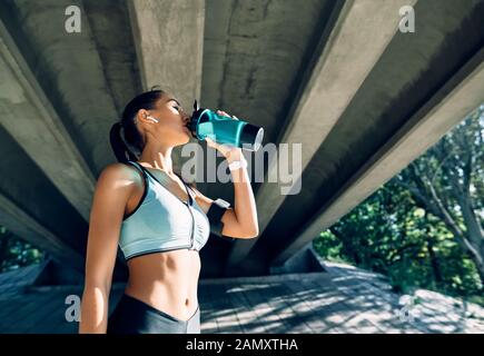 Jeune femme de fitness buvant de l'eau de la bouteille. Une femme sportive se reposant après une activité physique dans une ville industrielle à proximité de la rivière Banque D'Images