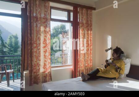 Mid adult woman Traveler d'Affaires indiennes de l'ethnicité en habits, portant sur un lit d'une villa de vacances et tenant une tasse de thé chaud dans mes rêves Banque D'Images