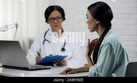 Une jeune femme qui dit des symptômes, un médecin expérimenté qui tient des dossiers médicaux Banque D'Images