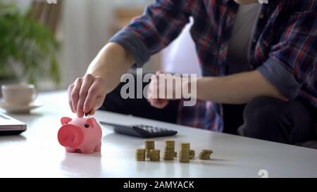 Jeune homme calculant de l'argent, mettant des pièces dans la banque piquante, budget familial Banque D'Images