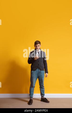 Choqué. Jeune homme caucasien utilisant smartphone, serfing, chat, Paris. Portrait complet isolé sur fond jaune. Concept des technologies modernes, des millénaires, des médias sociaux. Banque D'Images
