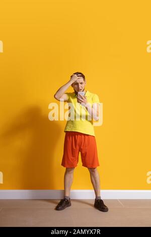 Choqué, étonné. Jeune homme caucasien utilisant smartphone, serfing, chat, Paris. Portrait complet isolé sur fond jaune. Concept des technologies modernes, des millénaires, des médias sociaux. Banque D'Images