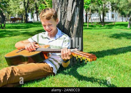 Un garçon avec une guitare sous un arbre, un adolescent joue et chante dans le parc. Banque D'Images