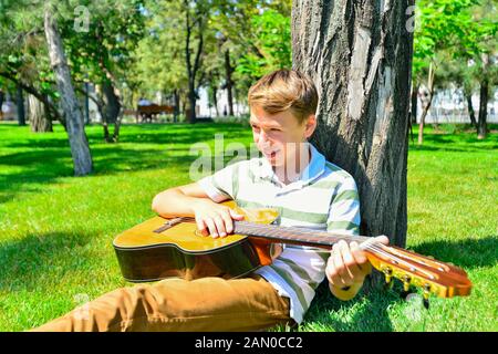 Un garçon avec une guitare sous un arbre, un adolescent joue et chante dans le parc. Banque D'Images