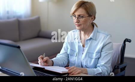Vieille femme en fauteuil roulant ayant cours en ligne avec l'enseignant DE L'INFORMATIQUE, prenant des notes Banque D'Images