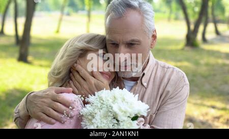 Old man hugging heureux femme tenant des fleurs, la célébration de mariage anniversaire Banque D'Images