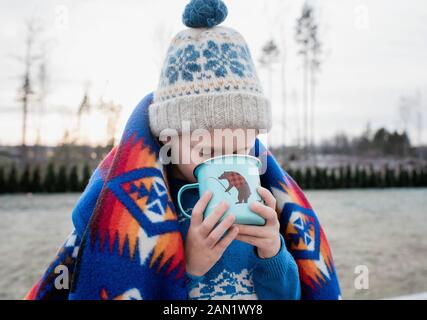 jeune garçon enveloppé dans une couverture buvant du chocolat chaud à l'extérieur Banque D'Images