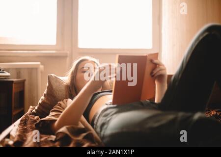 Teenage girl reading book while lying on bed at home Banque D'Images