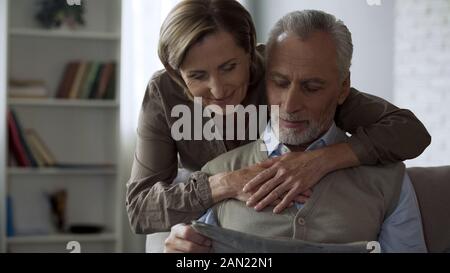 Vieillissement dame serrant son mari, homme assis sur la table et l'amour de journal de lecture Banque D'Images