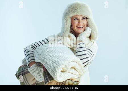 heureuse fille élégante, avec sweat à rayures blanches, écharpe et rabats d'oreille chapeau avec une pile de couvertures chaudes regardant l'espace de copie sur le dos bleu clair d'hiver Banque D'Images