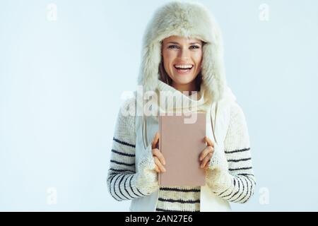 Portrait d'une jeune femme souriante de 40 ans en sweat à rayures blanches, écharpe et rabats d'oreille chapeau avec un livre sur fond bleu clair d'hiver. Banque D'Images