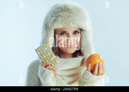 Portrait d'une jeune femme souriante de 40 ans en sweat-shirt blanc à rayures, écharpe et rabats d'oreille chapeau montrant un paquet orange et blister de vitamines isolées Banque D'Images