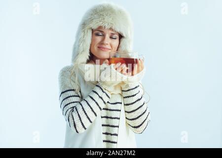 femme élégante et décontractée dans un pull à rayures blanches, un foulard et des rabats pour les oreilles chapeau, dégustant une tasse de thé chaud avec du citron et du miel sur fond bleu clair d'hiver. Banque D'Images