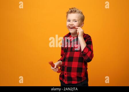 Le garçon mange des saucisses sèches. Beau garçon caucasien souriant dans la chemise rouge sur fond jaune. Horizontale, droite. Banque D'Images