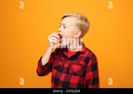 Le garçon mange des fruits, une alimentation saine pour les enfants. Beau garçon caucasien souriant dans la chemise rouge sur fond jaune. Horizontale, droite. Banque D'Images